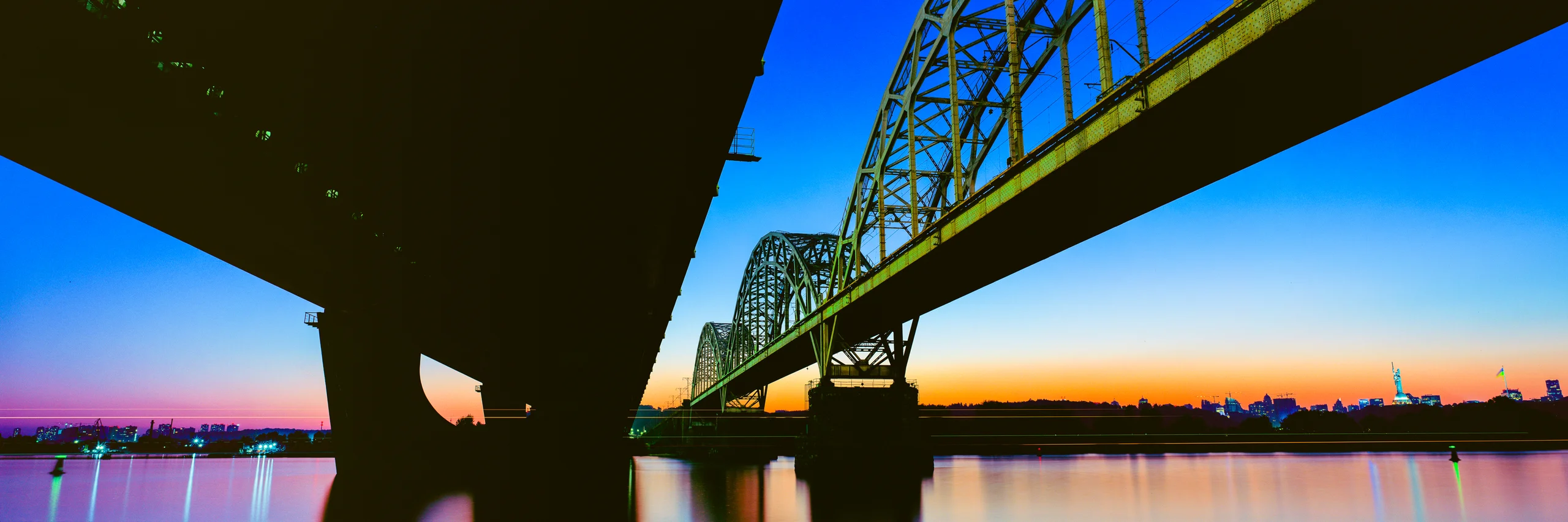 Panoramic fine art photograph of two bridges over a river at sunset, with a vivid blue and orange sky and city lights reflected in the water.