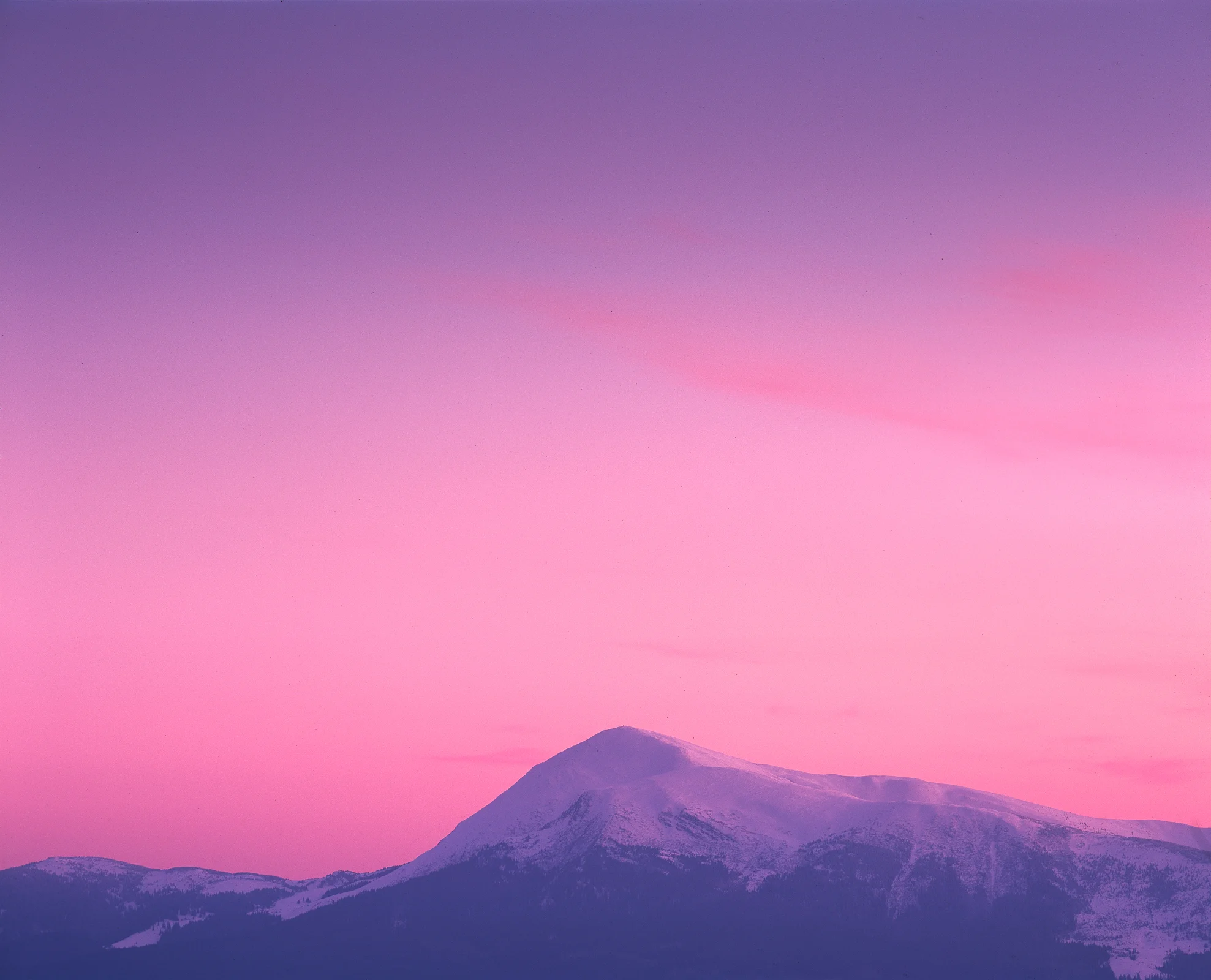 Snow-covered mountain ridge and dark wooded slopes beneath an expansive pink and violet sky.