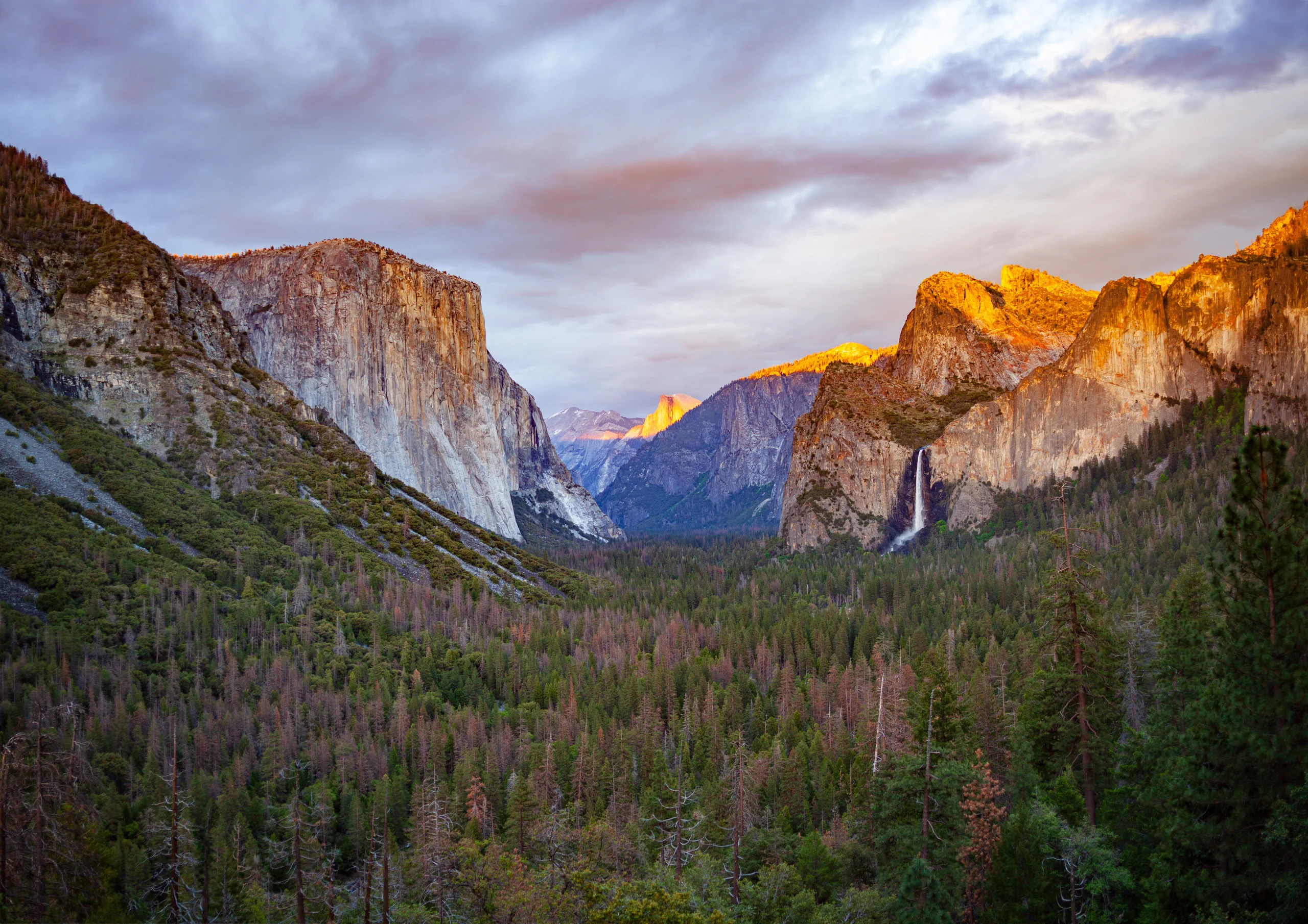 Forested mountain valley between granite cliffs, with golden light on the upper rock faces and a waterfall beneath a cloudy sky.