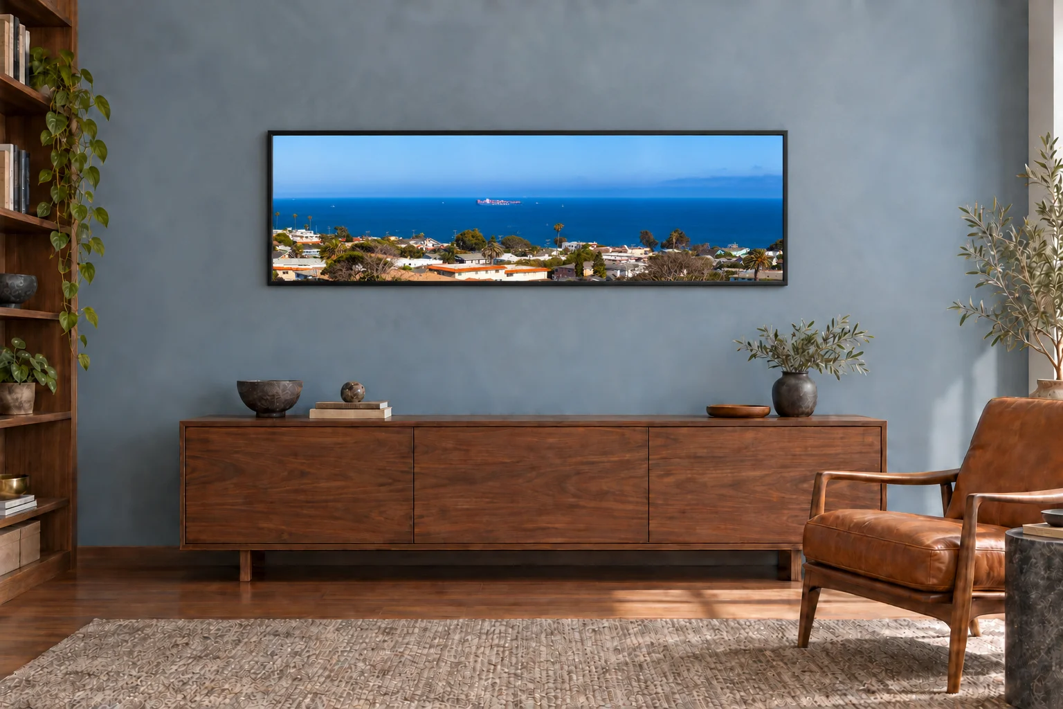 Illustrative mockup: a framed Los Angeles coastal panorama above a walnut credenza in a blue-grey study with a cognac leather chair.