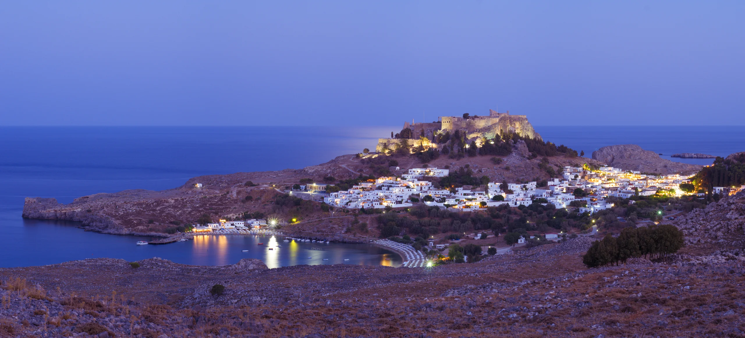 Coastal white village below an illuminated hilltop fortress, with a curved bay, reflected lights and blue sea at dusk.