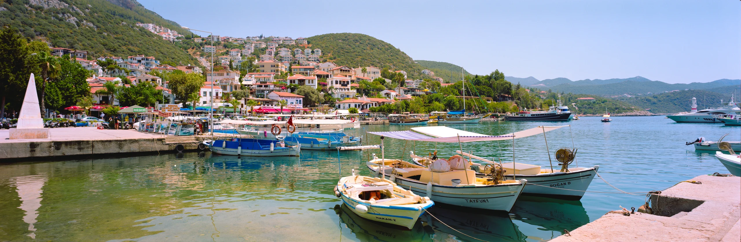 Moored boats beneath canvas shades in a sunny harbour with hillside houses and green water.