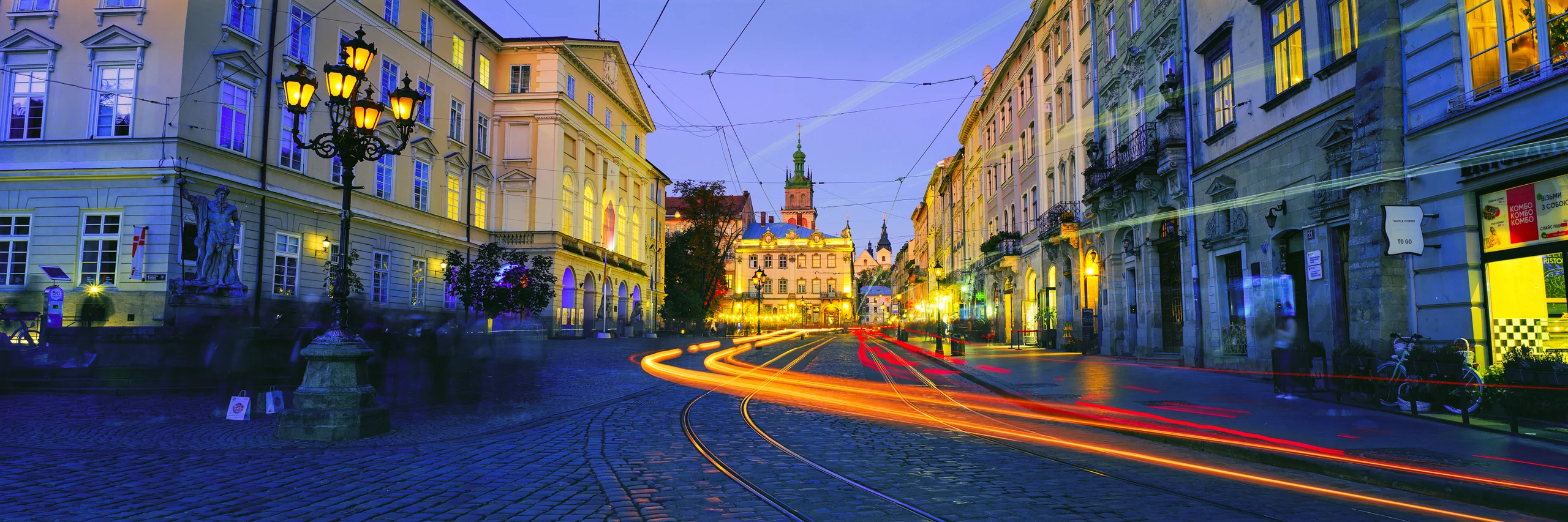 Golden and red light trails curve along tram tracks between historic buildings and glowing street lamps at blue hour.
