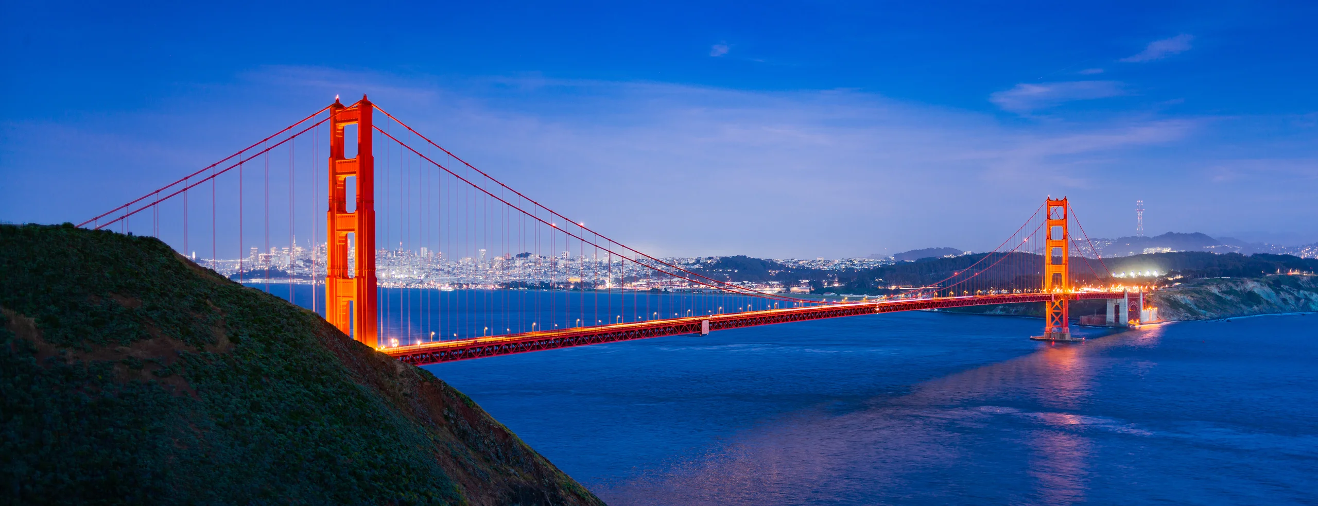 Illuminated Golden Gate Bridge above blue water at dusk, with a dark headland in the foreground and city lights beyond.