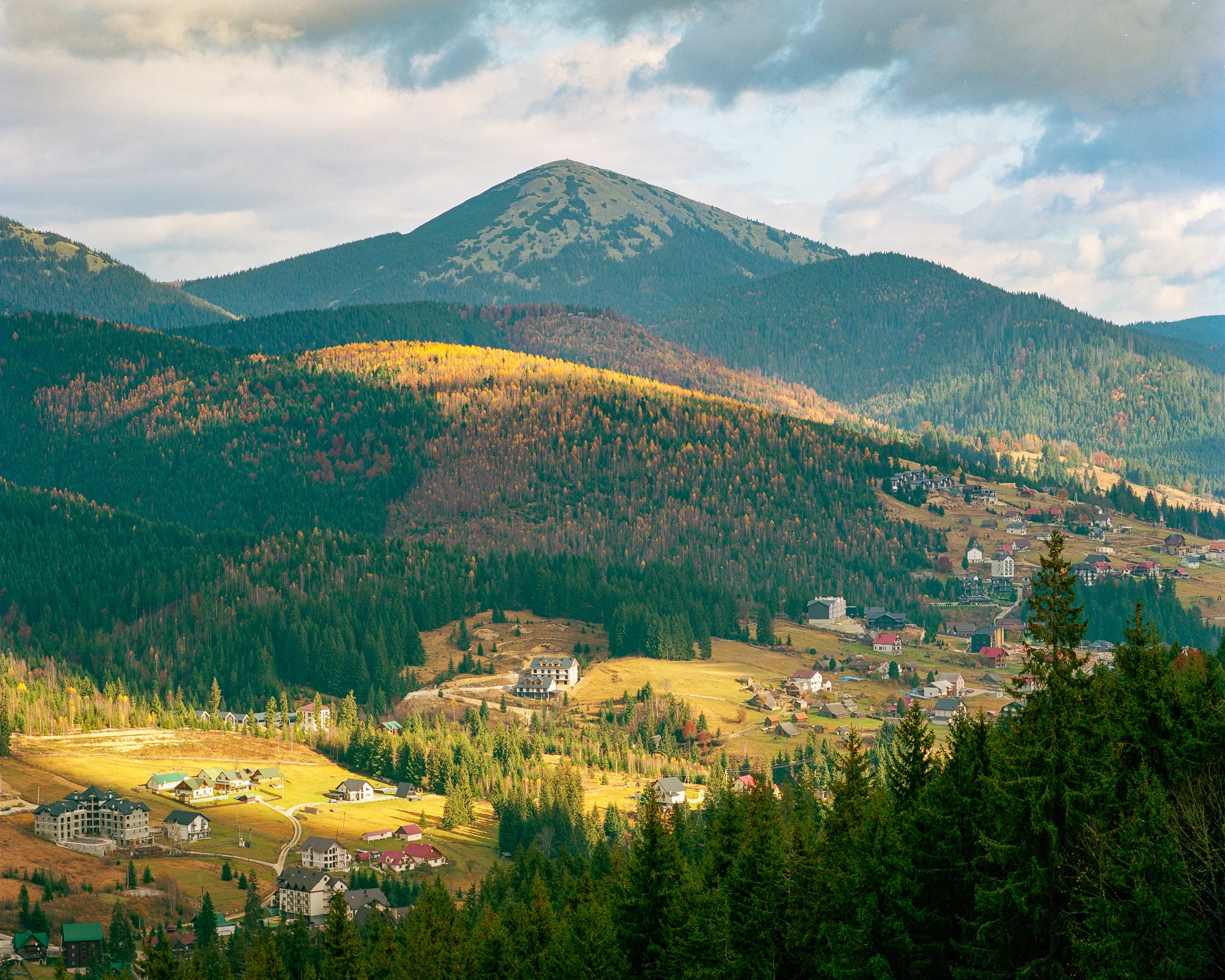 Autumn mountain village with sunlit meadows, golden trees, dark firs and a rounded forested summit beneath clouds.