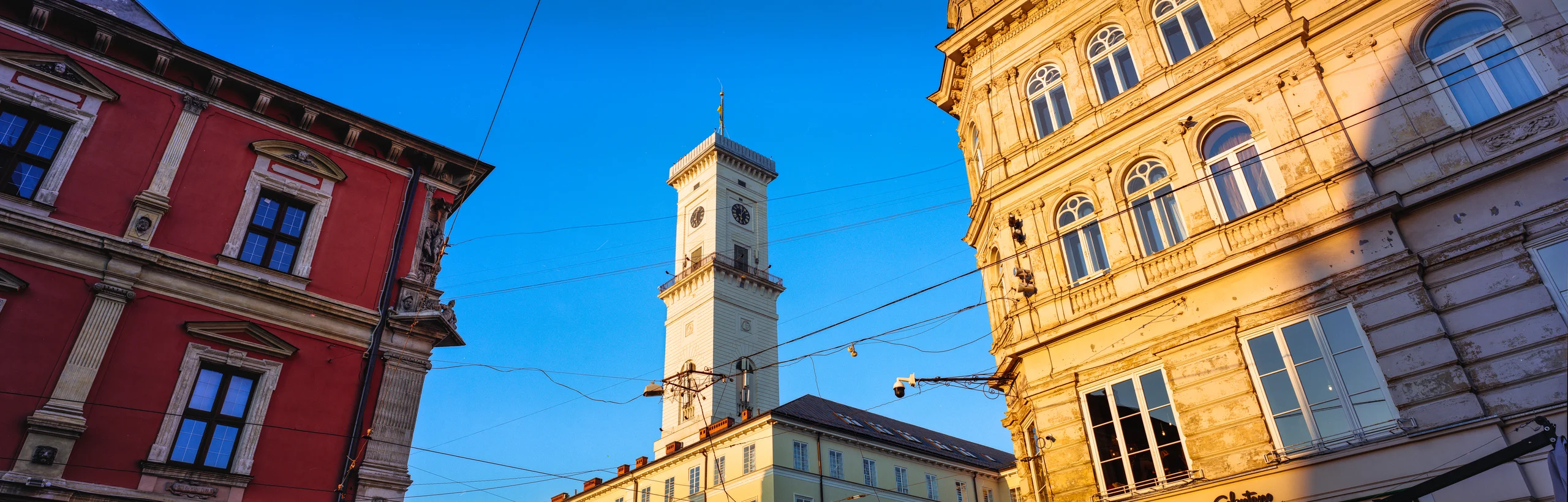 Lviv City Hall clock tower between red and sunlit golden façades, with overhead wires against blue sky, Ukraine.