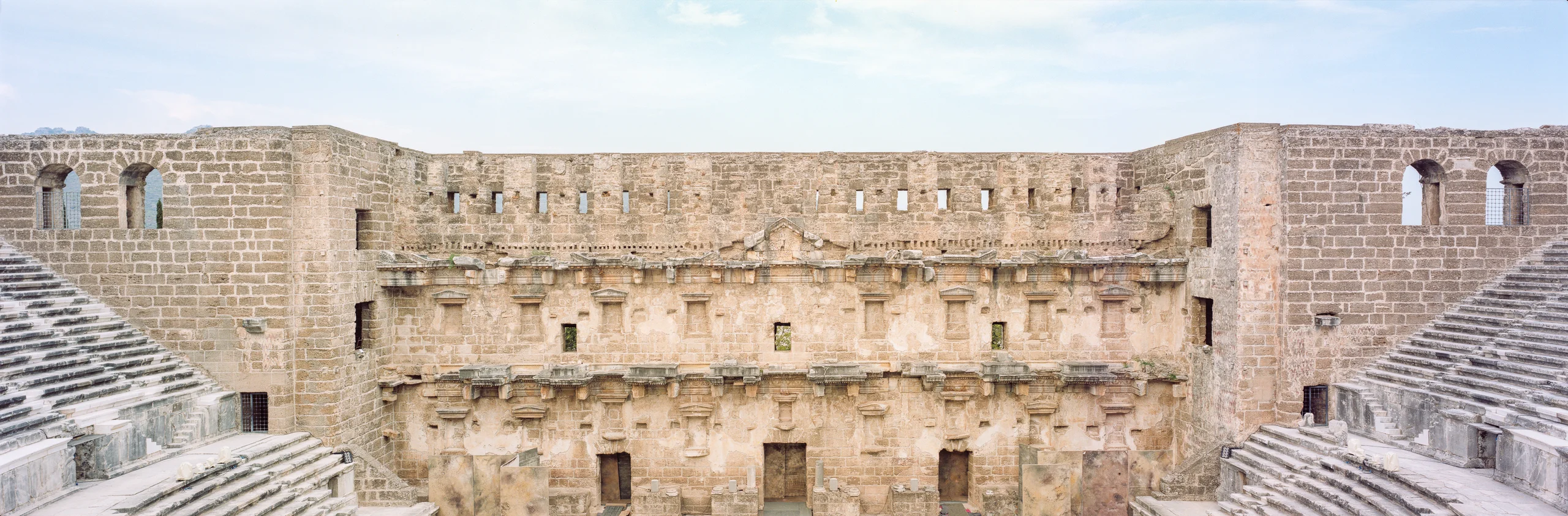 Panoramic view of an empty stone theatre, with stepped seating on both sides and an ornate stage wall beneath a pale blue sky.