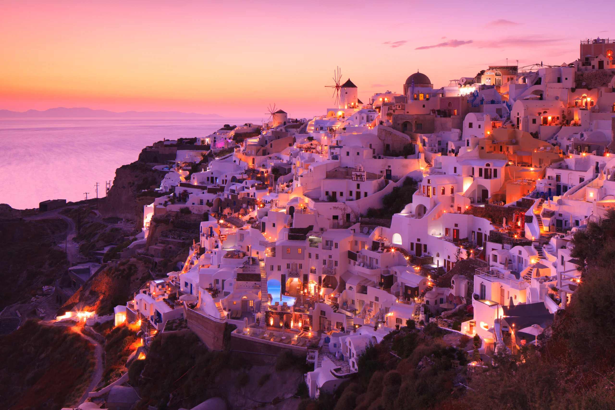 Whitewashed Santorini buildings, domes and windmills glow above the Aegean beneath a pink and peach evening sky.