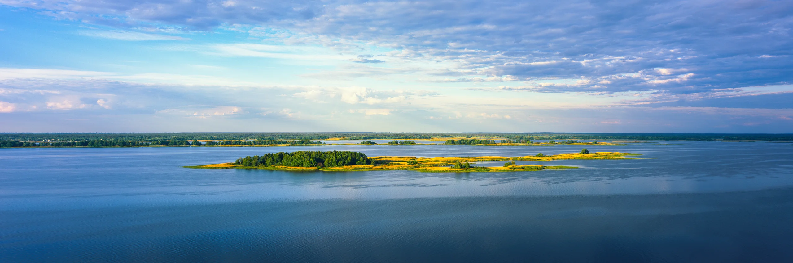 Panorama of blue water surrounding low islands with green trees and sunlit golden grasses beneath a wide, cloud-filled sky.