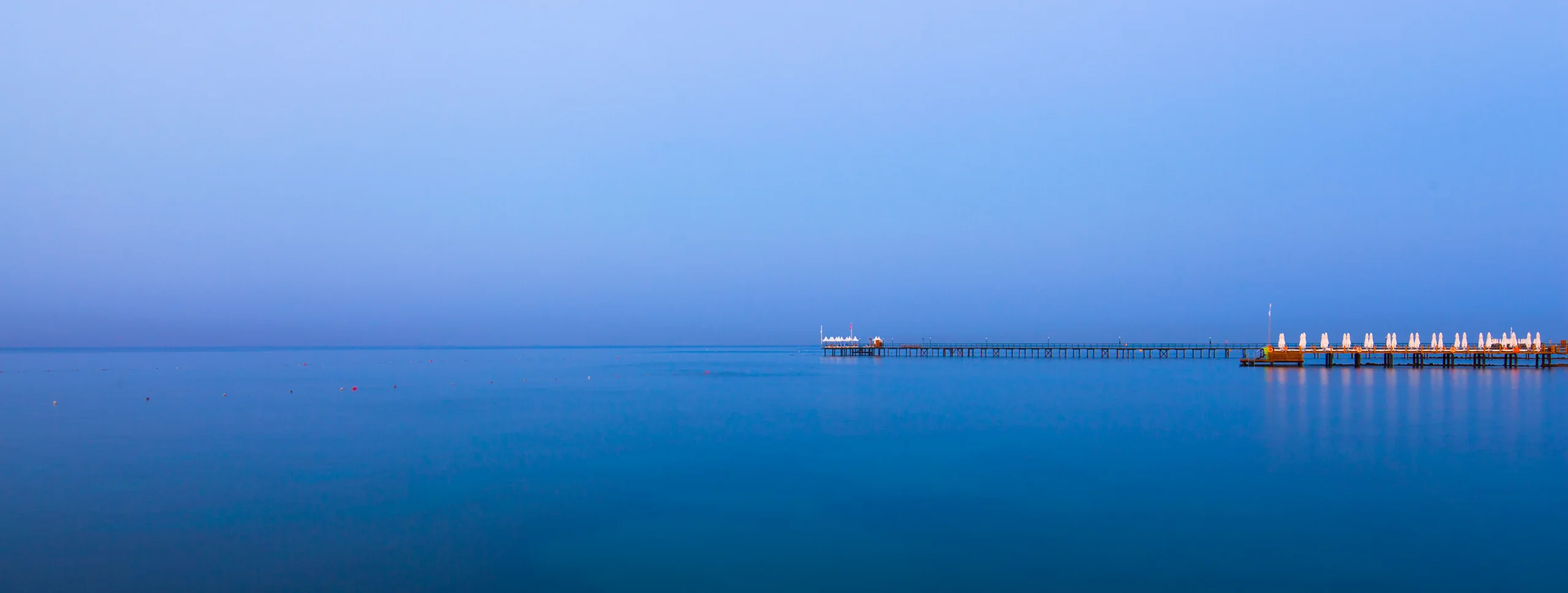 Still blue sea beneath a pale sky, with a long pier and rows of white umbrellas on the right.