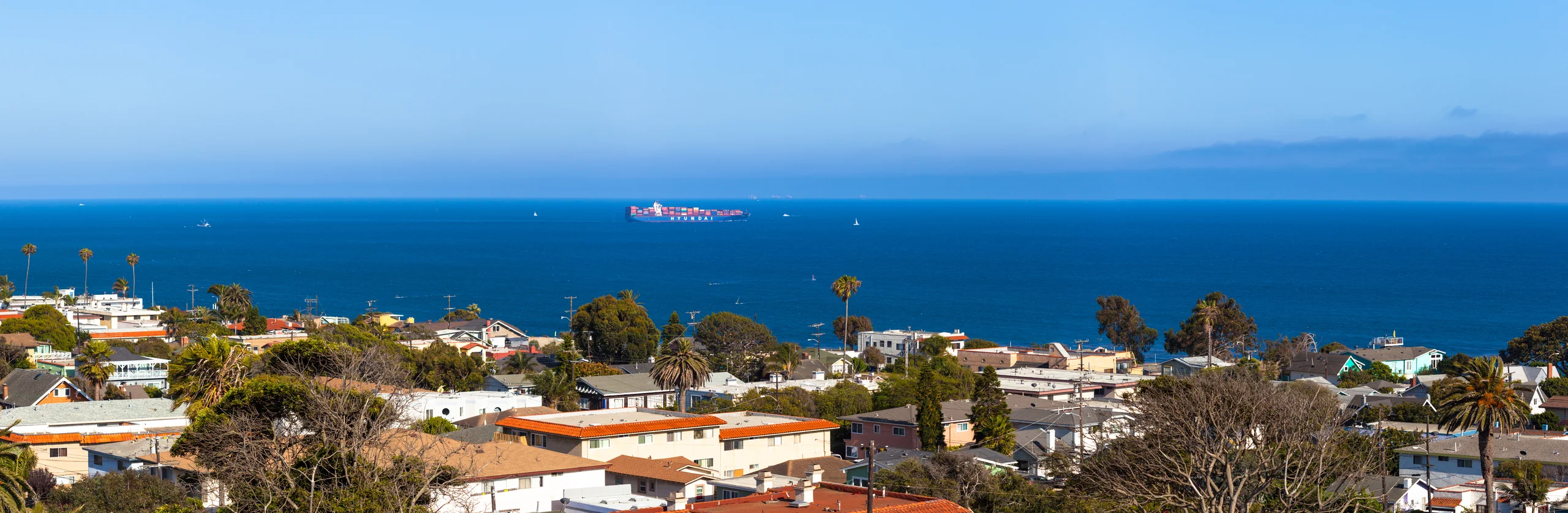 Panorama of Los Angeles rooftops and palms overlooking a blue ocean, with a distant cargo ship and small sailboats.