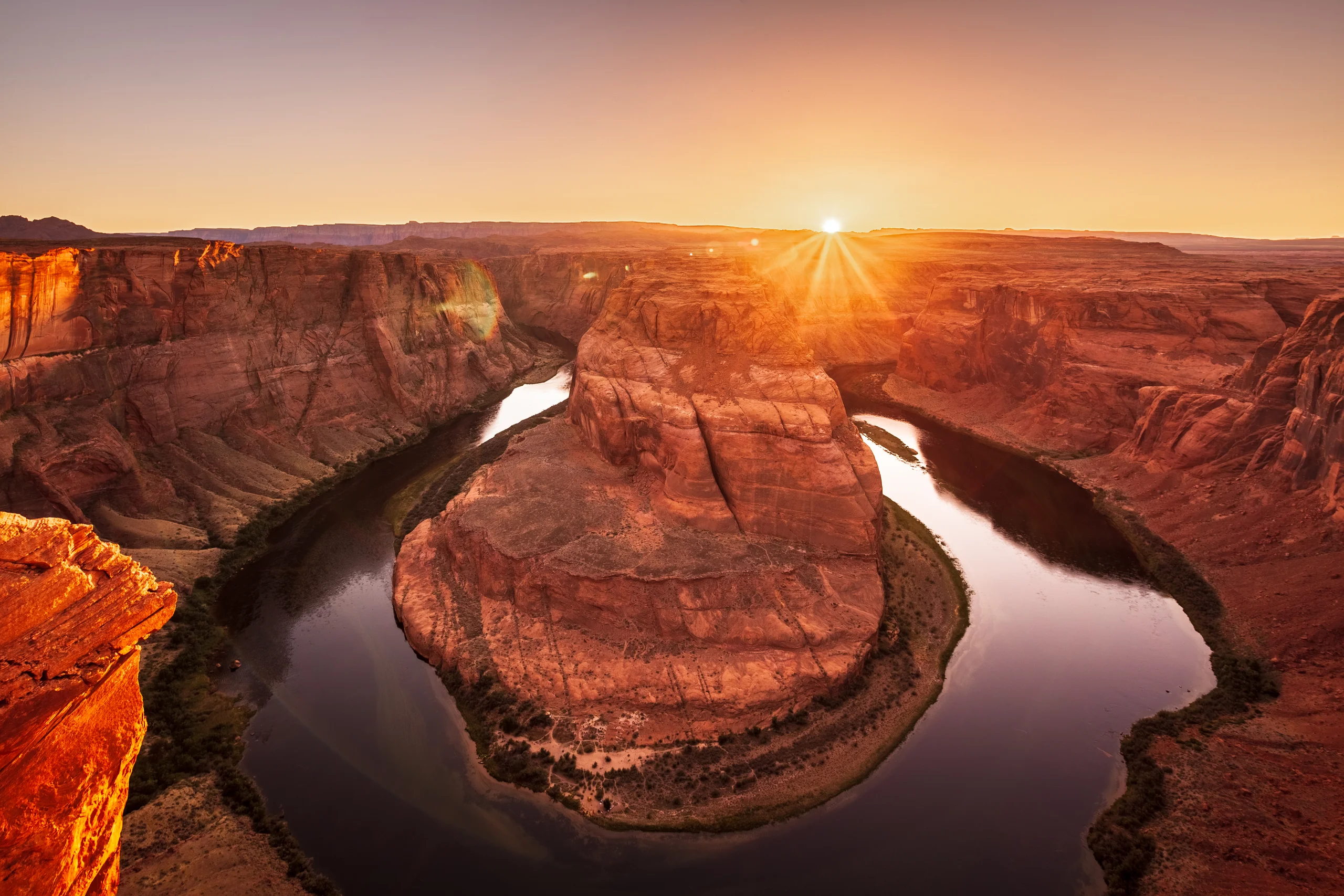A river bending around red sandstone cliffs beneath a low sun and amber sky.