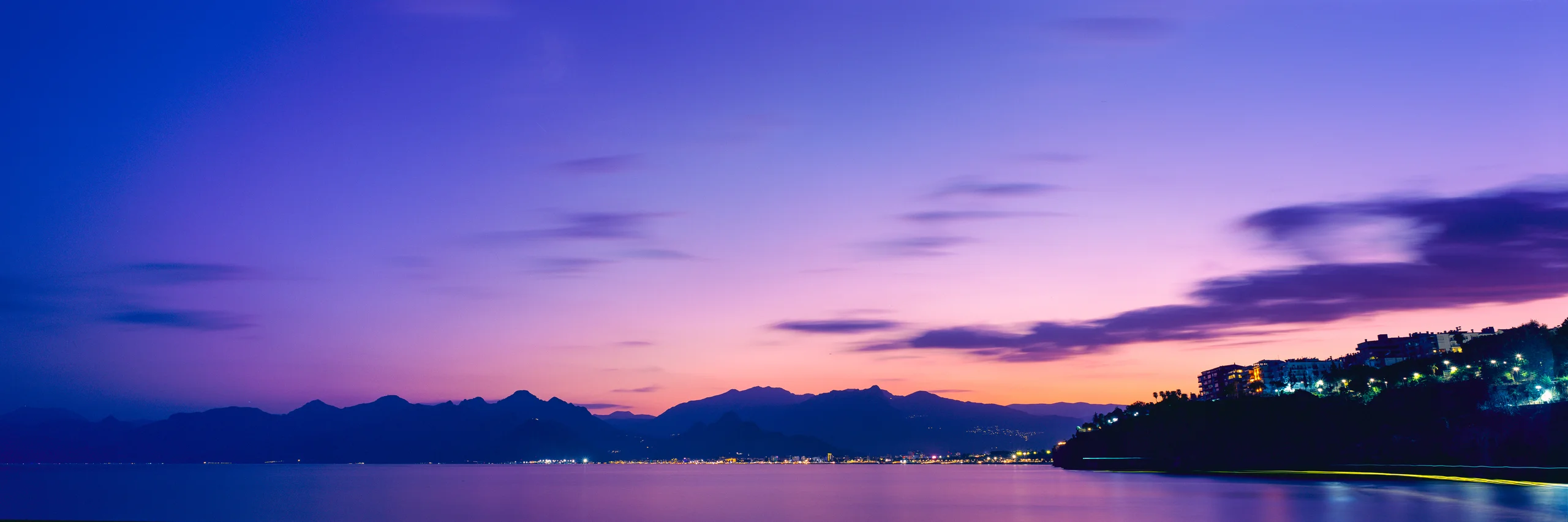 Panoramic bay beneath a rose, violet and blue sky, with dark mountain silhouettes, shoreline lights and illuminated buildings on the right headland.