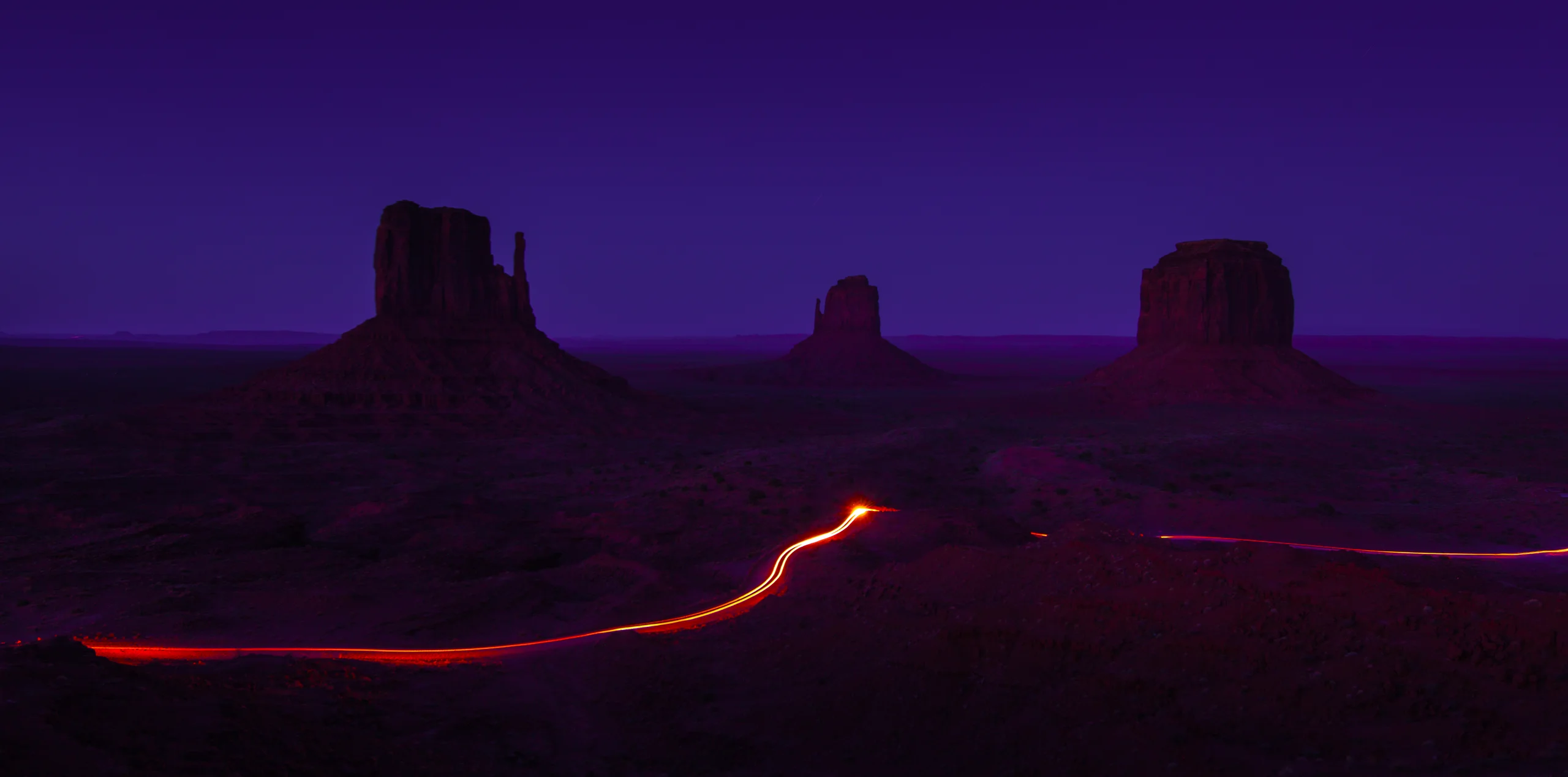 Three desert rock formations beneath a violet sky, with a curving orange-red vehicle light trail in the foreground.