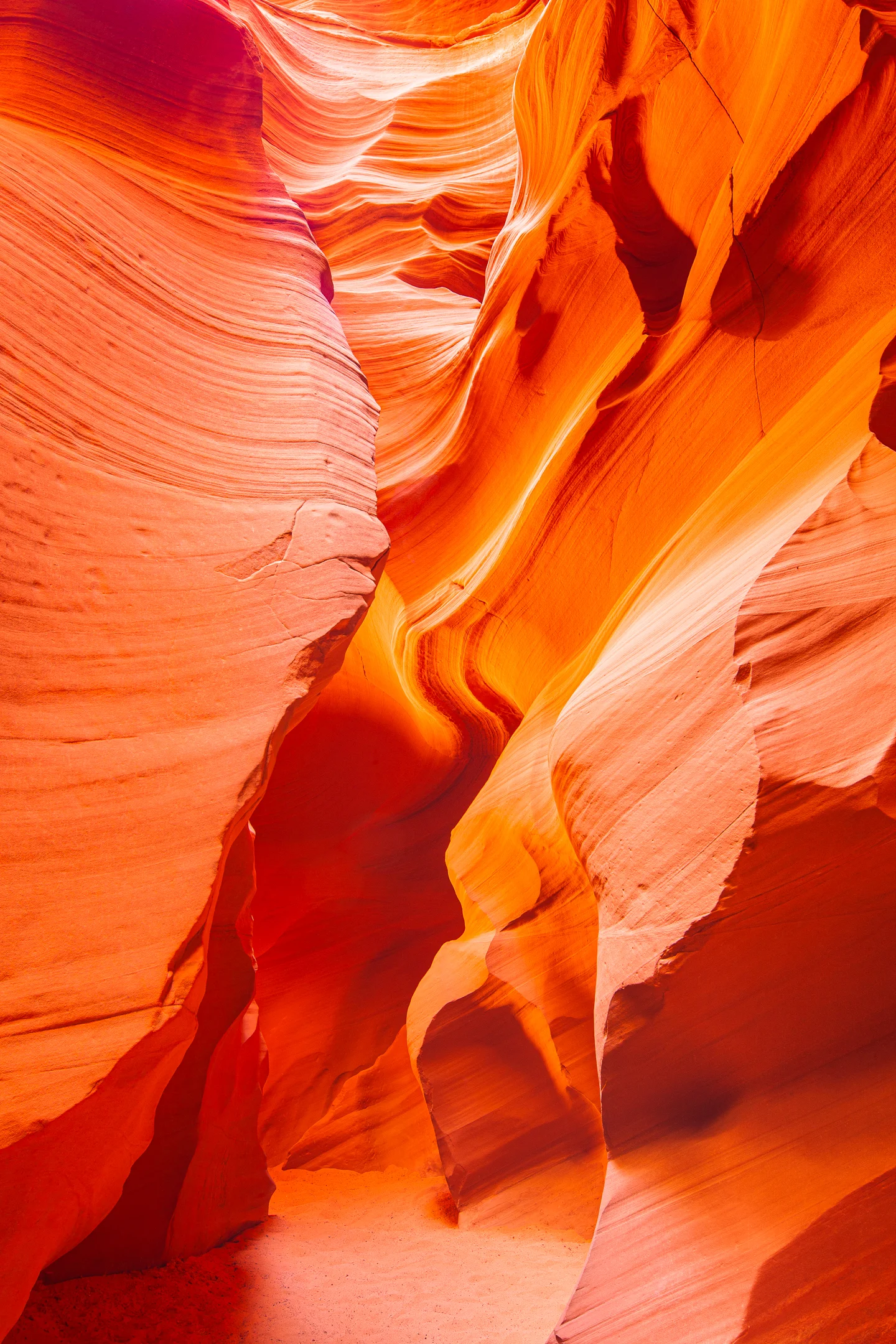 Glowing orange and red sandstone walls curve around a narrow sandy canyon passage, their surfaces marked by fine flowing layers.