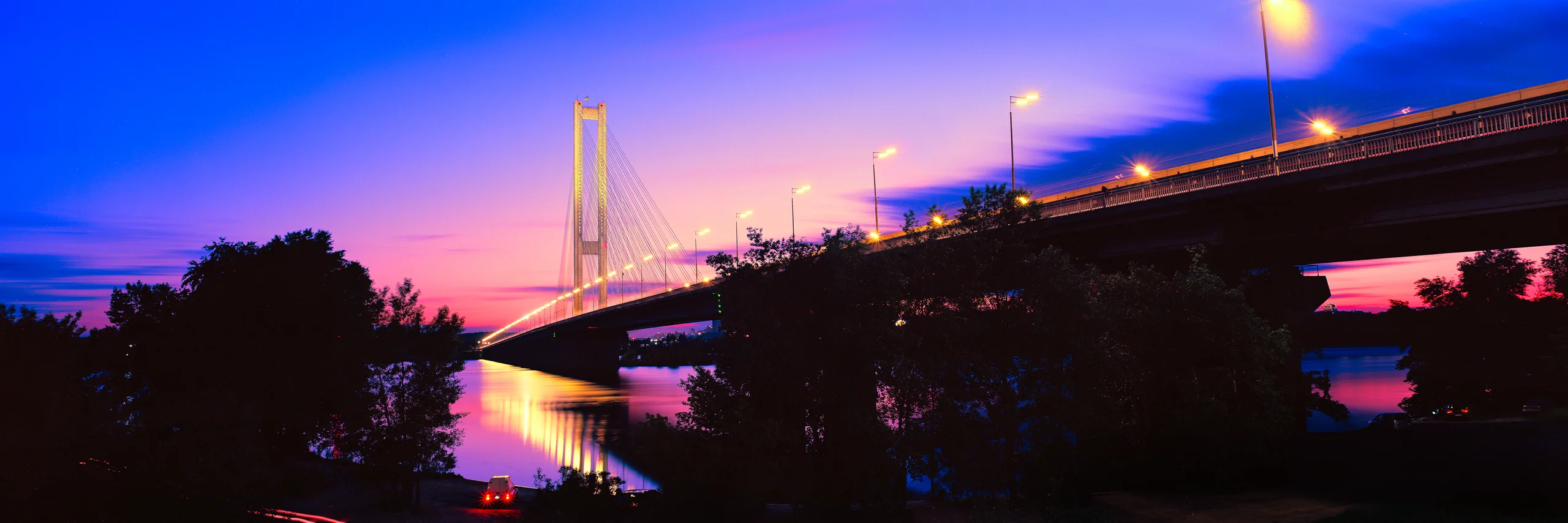 Panoramic evening view of a bridge over the Dnipro in Kyiv, with a pink and violet sky, illuminated structure and city lights reflected in the water.