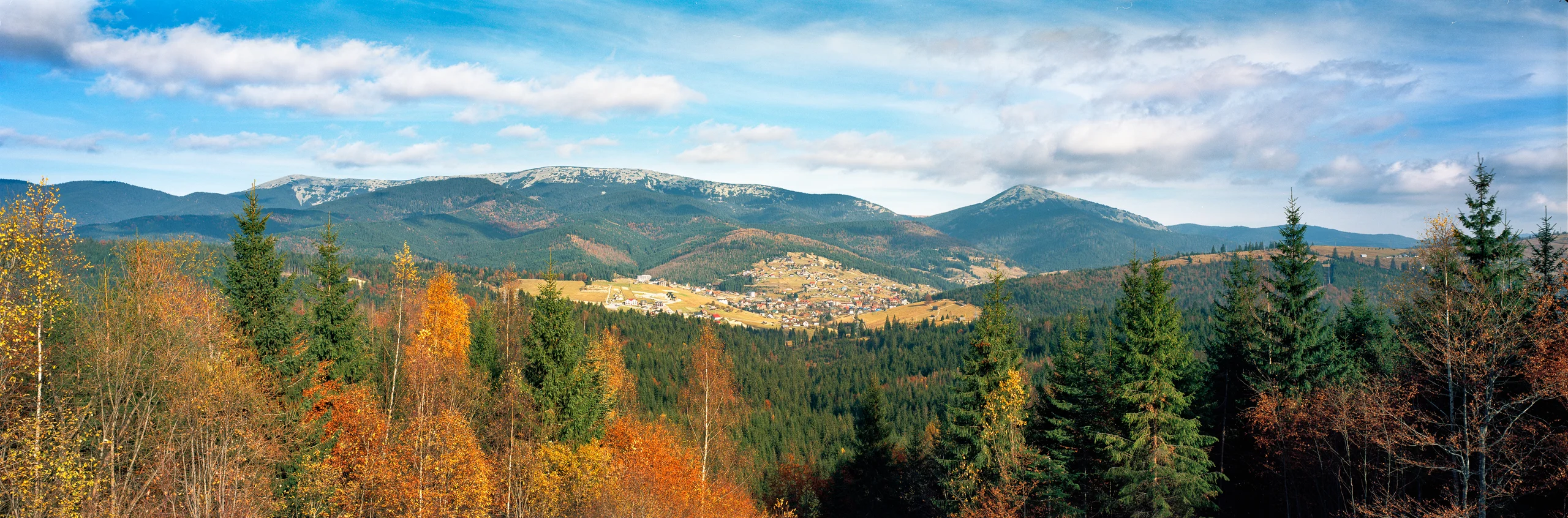 Autumn panorama of golden and rust-coloured trees, dark conifers, a village and fields beneath forested mountain ridges and a cloudy blue sky.