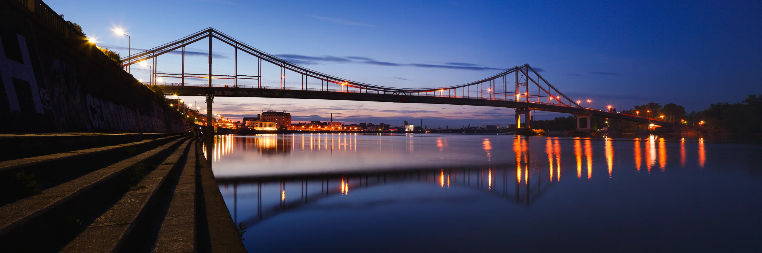 Panoramic suspension bridge at dusk, with amber lights reflected in blue water and stone embankment steps in the foreground.