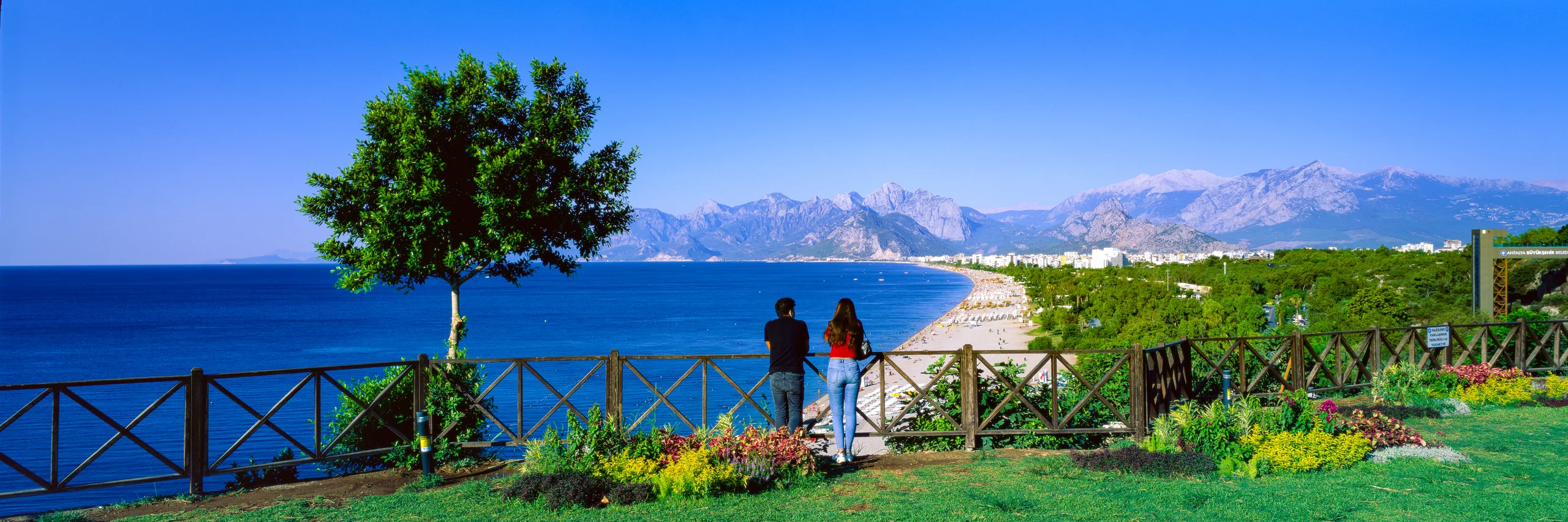 Two people at a garden railing overlooking a blue sea, curving beach, distant city and mountains.
