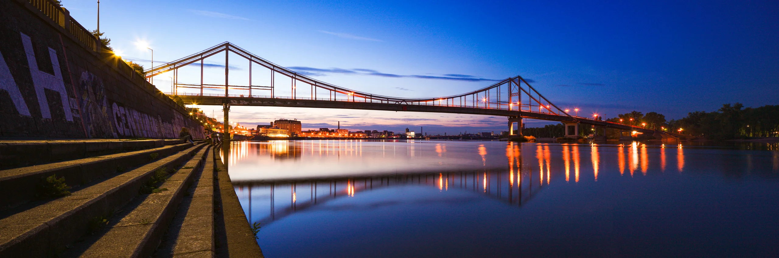 Panoramic suspension bridge at dusk, with amber lights reflected in blue water and stone embankment steps in the foreground.