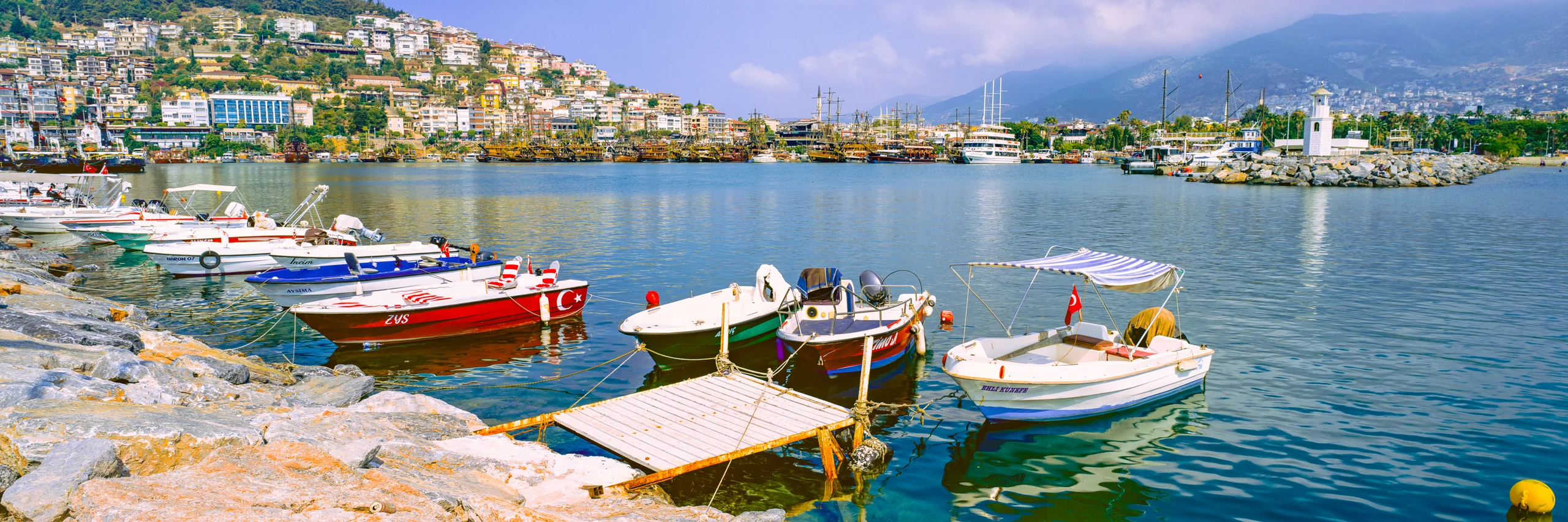 Colourful small boats and a wooden landing in Alanya harbour, with blue-green water, hillside houses, a white lighthouse and distant mountains.