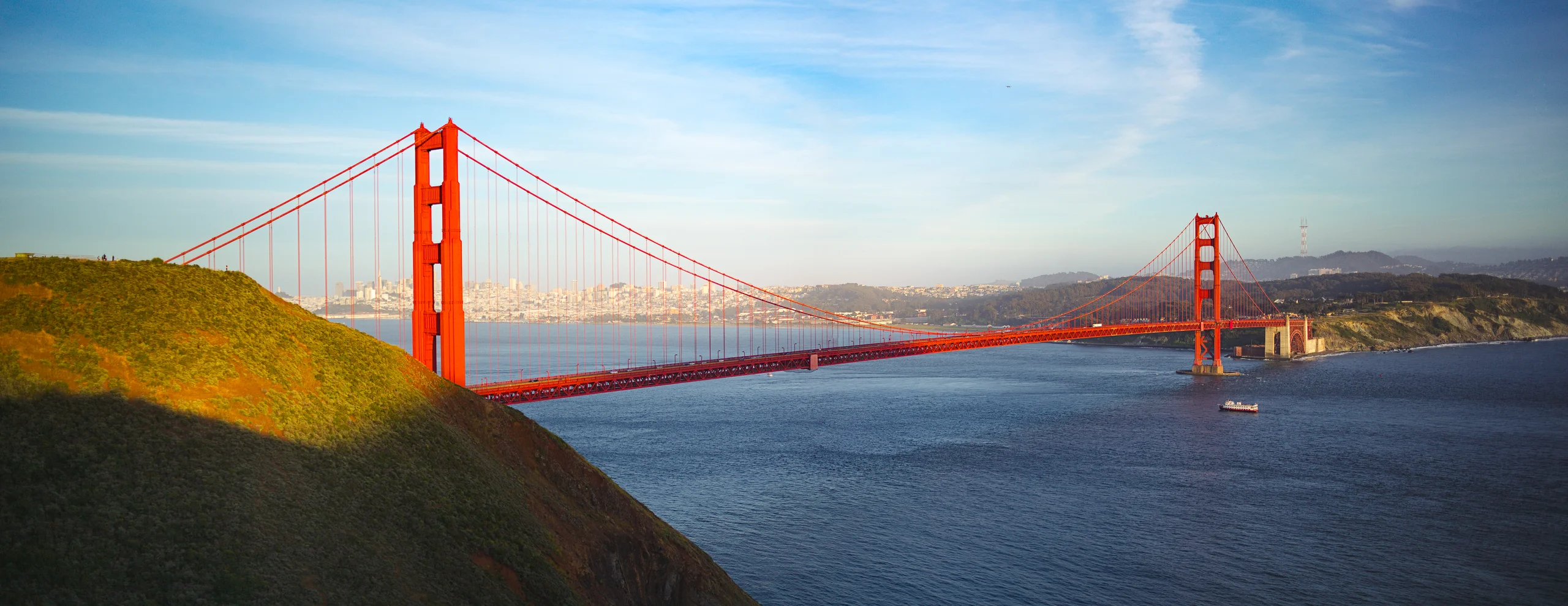 Golden Gate Bridge spanning blue water, with red towers and suspension cables, a sunlit foreground hillside and a distant city.