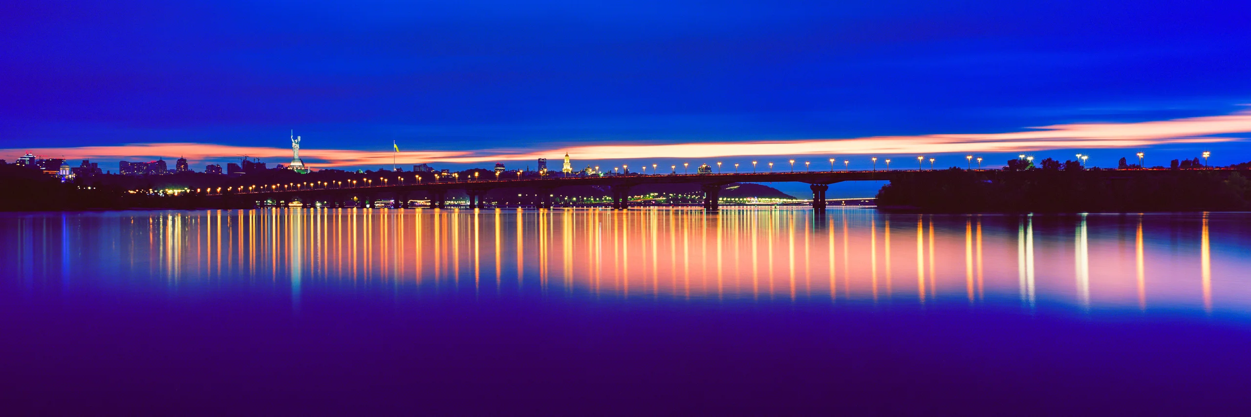 Kyiv at dusk, with city lights and an illuminated bridge viewed from the beach.
